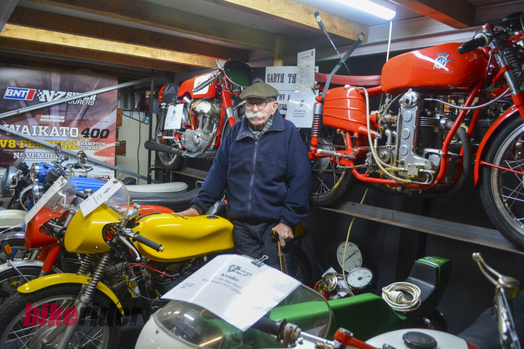 Older man wearing a flat cap and glasses stands among bright red vintage motorcycles in a workshop display.
