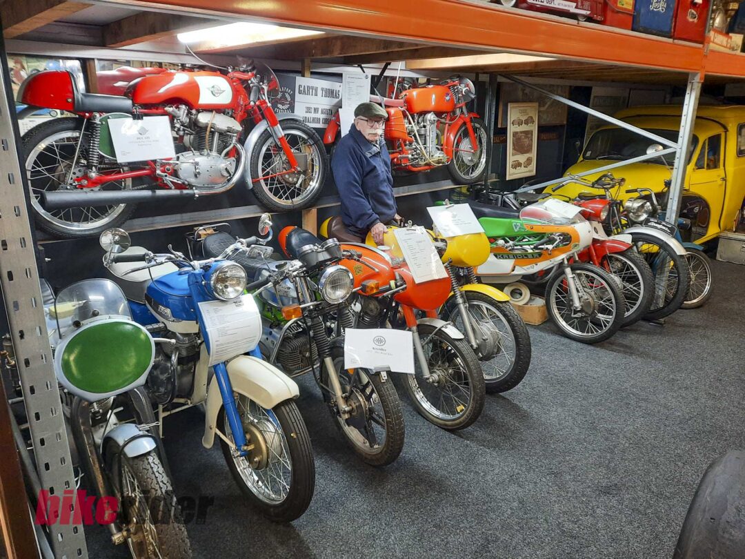 Elderly man in a cap standing beside a row of vintage red motorcycles on display in a showroom or museum setting.