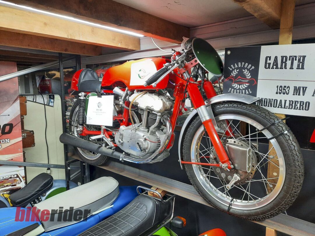 Red vintage motorcycle mounted on a wall display in a showroom, with other bikes and promotional signage in the background.