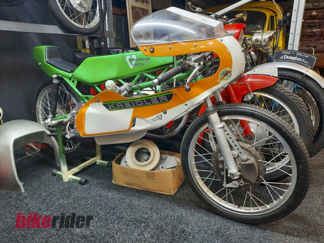 Vintage Kreidler racing motorcycle in orange, white and green livery on display in a museum or showroom, with another bike in the background.