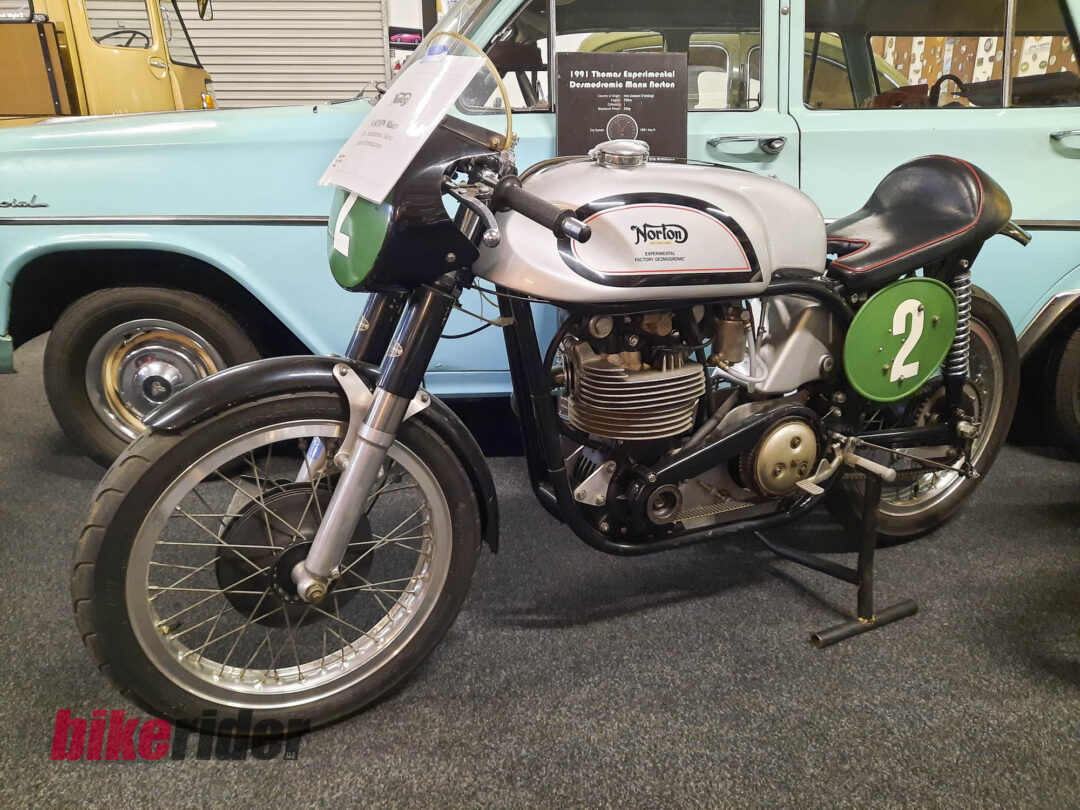 Vintage Norton motorcycle with a green racing plate marked '2' on a showroom floor, a light blue classic car visible in the background.