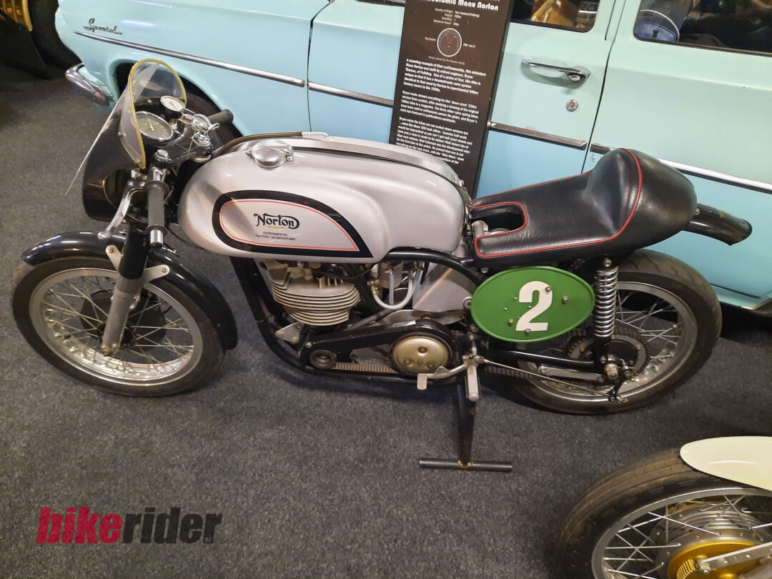 Vintage Norton motorcycle on display with a light blue car in the background; visible 'Norton' logo and a green number-2 plate on the side panel.