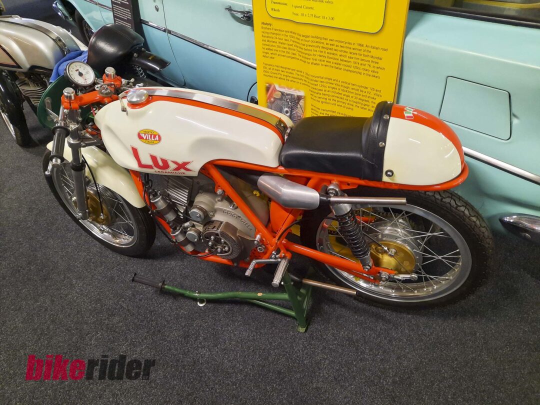 Vintage Moto Villa motorcycle on display, orange frame and cream body, with a teal bus and yellow information panel in the background.
