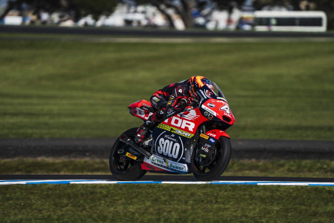 Motorcycle racer in red and black gear leaning into a high-speed turn on a race track, helmet bright orange and sponsor logos visible on the bike and suit.