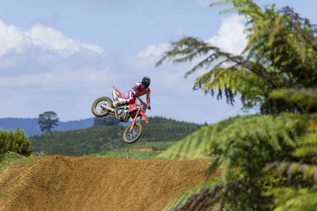 Motocross rider in red gear leaping mid-air from a dirt jump against a blue sky and green trees.