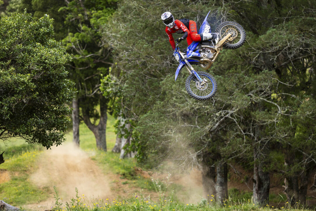Motocross rider in red gear mid-air on a blue dirt bike over a forest trail.