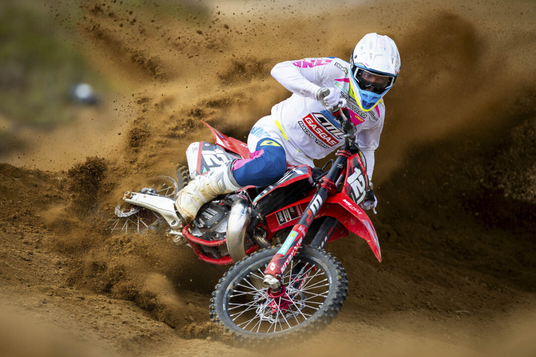 Motocross rider in white gear on a red bike carving through dirt at high speed, kicking up a cloud of soil.