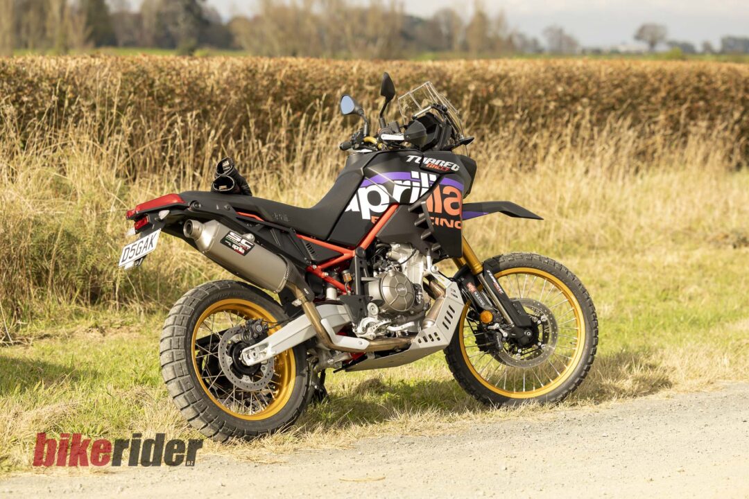 Side view of a black, purple, and orange rally-style motorcycle parked on a dirt path in a grassy field with tall brush and distant trees; 'bikerider' text is visible on the ground in the foreground.