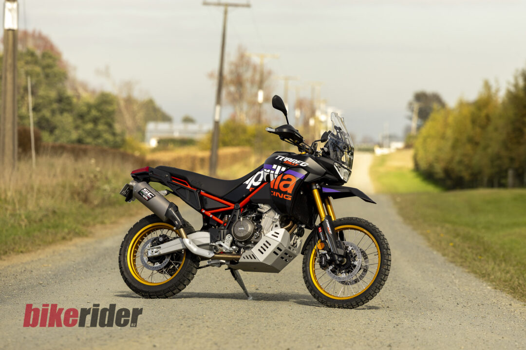 Side view of a black and orange sports motorcycle parked on a rural road, with a clear windscreen and yellow rims in focus against a blurred background of vegetation.