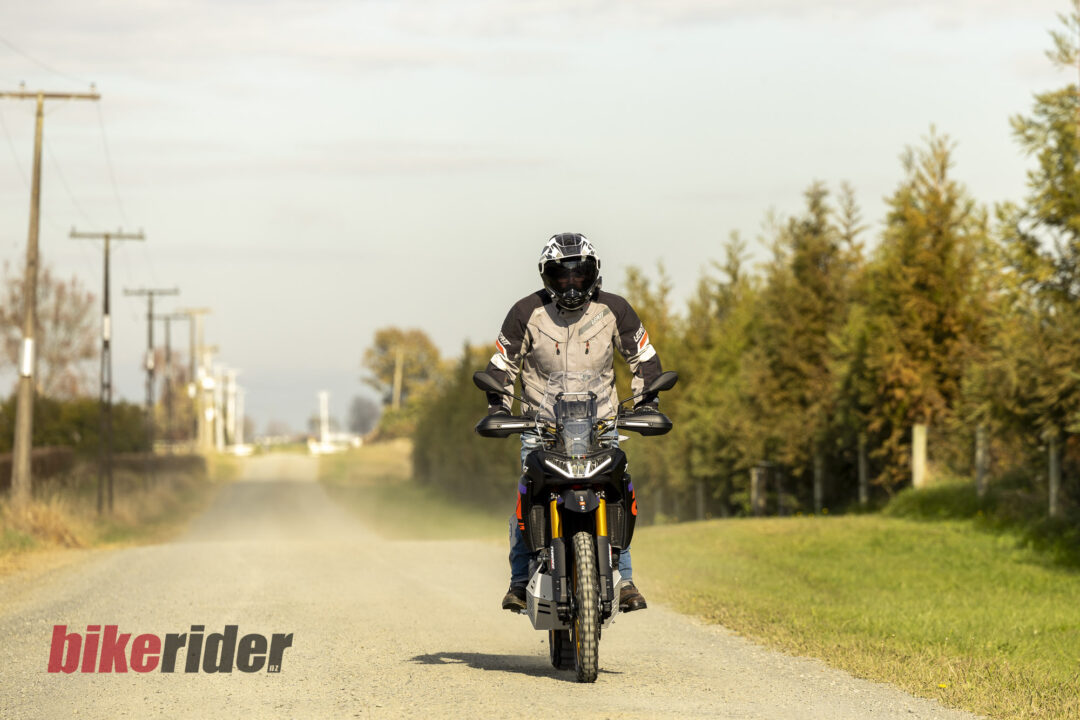 Motorcycle rider wearing a helmet and protective gear riding toward the camera on a rural road lined with trees.