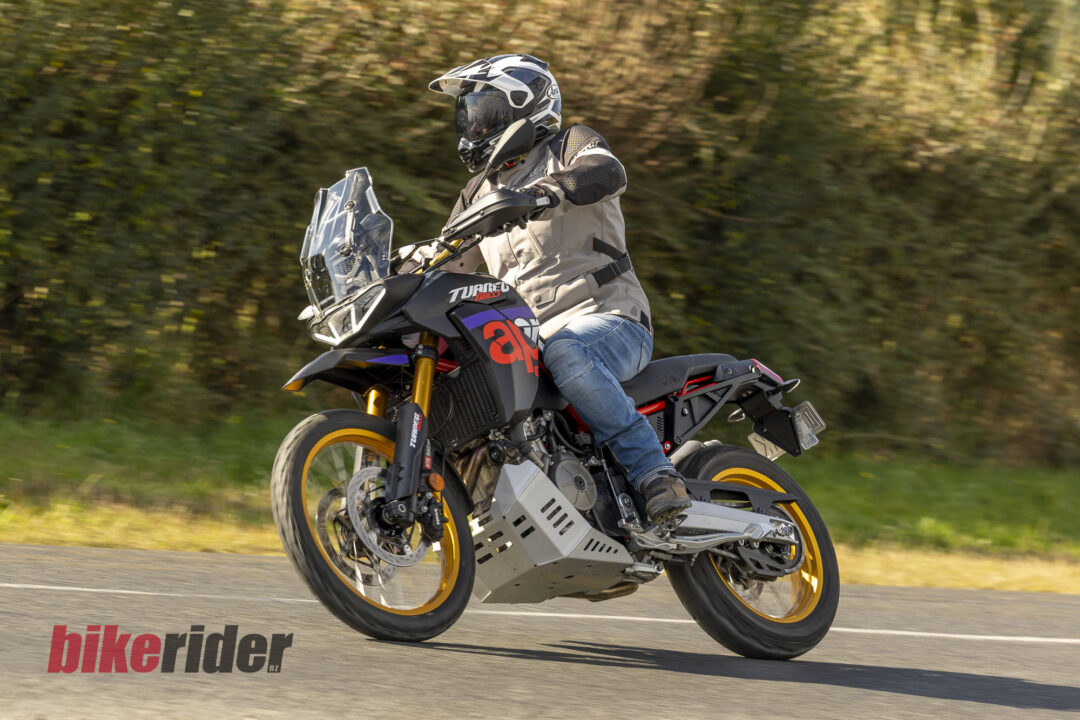 Motocycle rider wearing protective gear leans into a turn on a road, riding an adventure bike with orange wheels, blurred background, watermark ‘bikerider.nl’ in the corner.