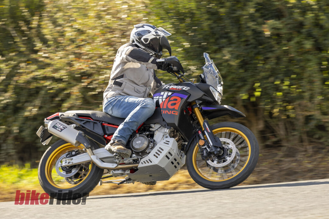 Motorcyclist wearing a full-face helmet and beige jacket leans into a turn on a black/orange sport-touring bike, blurred green background behind.