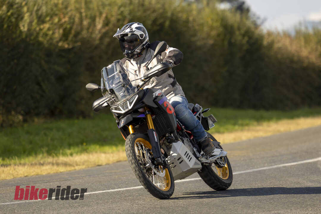 Motorcyclist wearing protective gear and helmet leans into a left turn on a dual-sport bike, riding on a paved road with a tree-lined background.