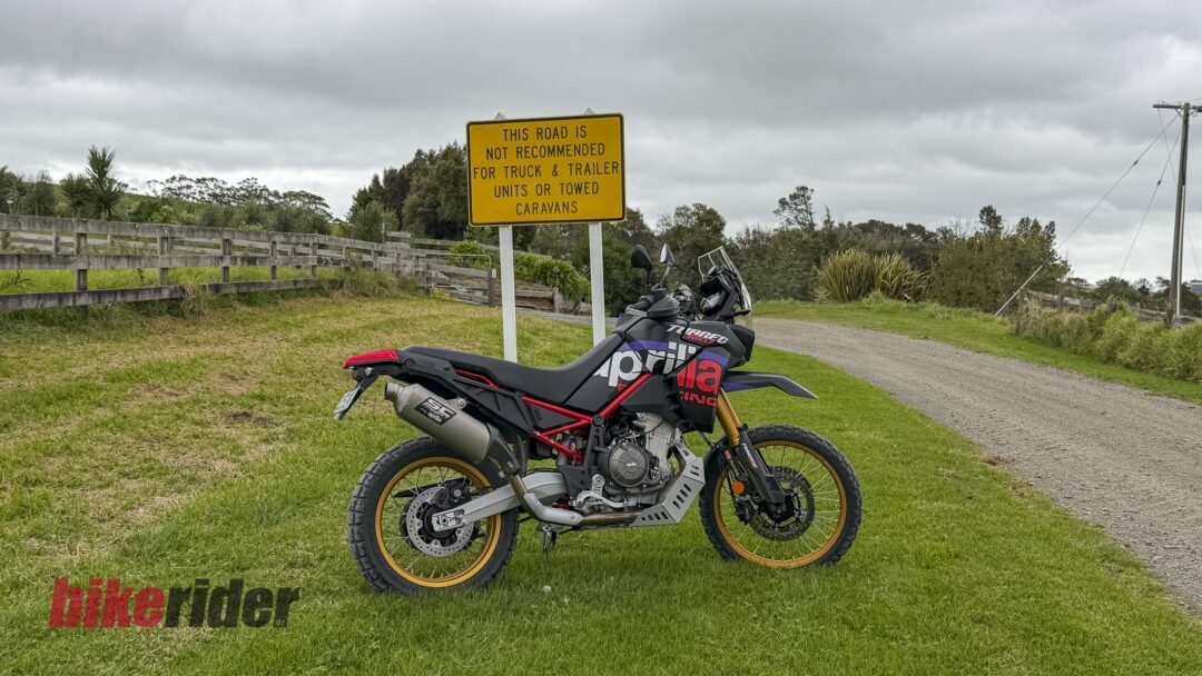 Black and purple Aprilia adventure motorcycle parked on grass beside a yellow warning sign in a rural area with a cloudy sky.