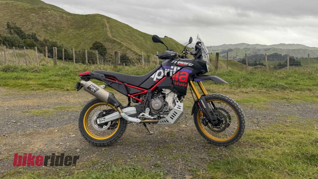 Adventure motorcycle parked on a gravel road with green hills and a wooden fence in the background under a cloudy sky.