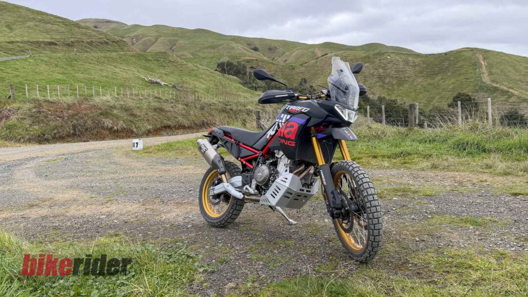 Adventure motorcycle parked on a gravel road with green rolling hills and a wooden fence in the background, under a cloudy sky.