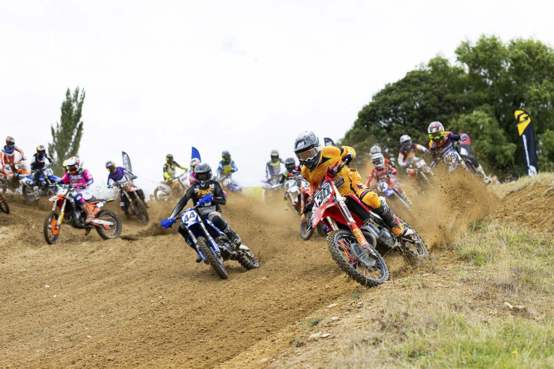 Motocross race: riders in colorful gear speeding around a dirt track, kicking up dust as they bend into a turn on an open outdoor course