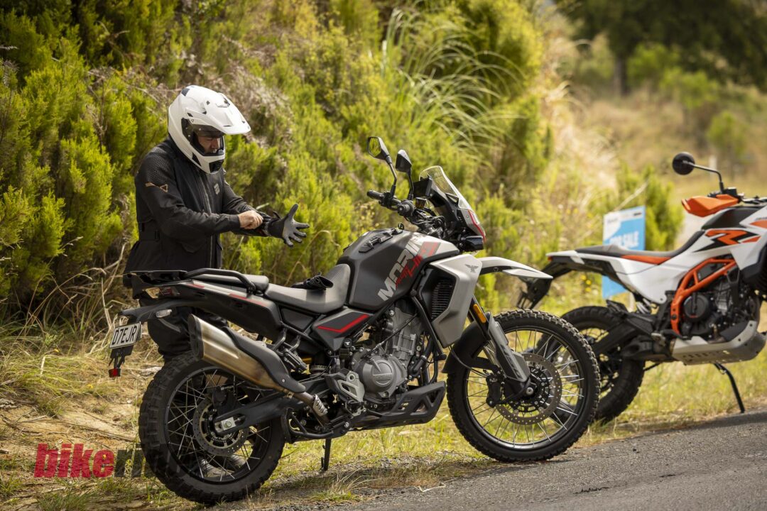 Motorcyclist in a white helmet adjusting gloves beside a black adventure motorcycle on a grassy roadside with bushes behind.
