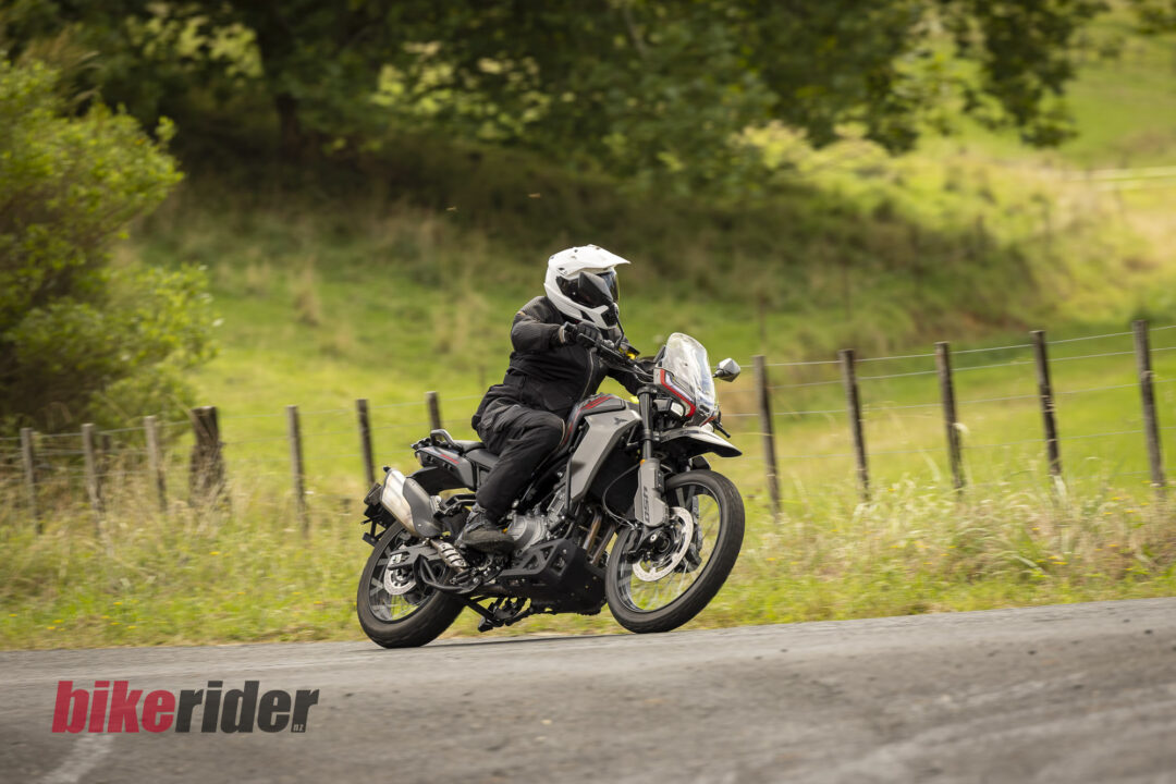 Motorcyclist in a white helmet riding a black adventure bike along a rural road with a green field and fence in the background.