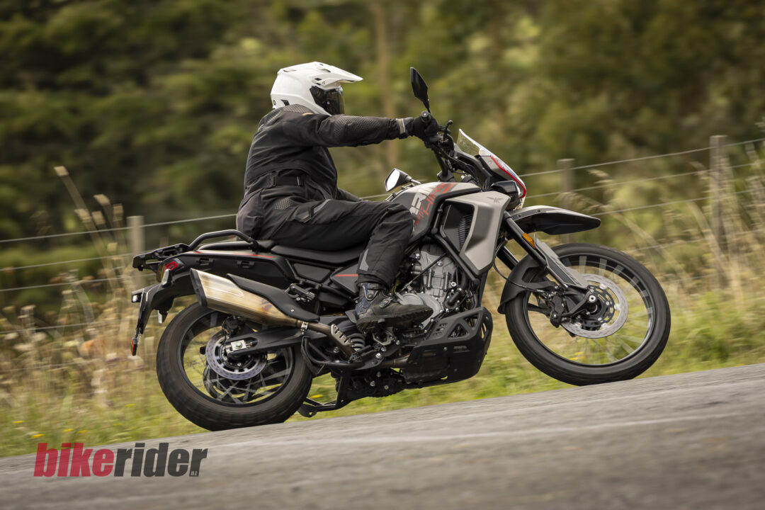Motorcyclist wearing a white helmet and black gear riding a grey adventure motorcycle on a rural road, logo visible on the road edge.
