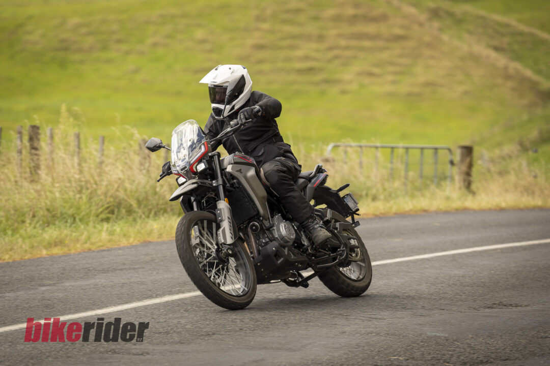 Motorcyclist in white full-face helmet and black protective gear leaning into a turn on a rural road with grassy hillside behind.