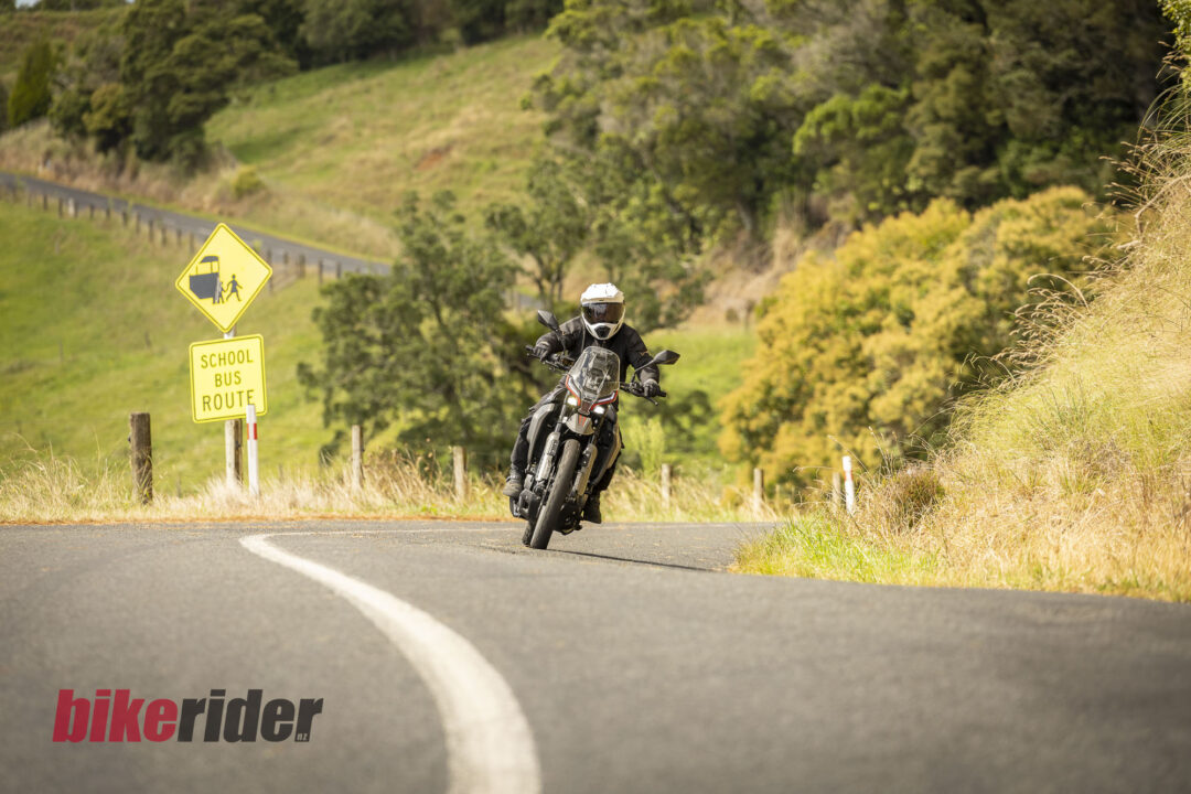 Motorcyclist wearing a white helmet and protective gear leans into a curve on a rural road, with a yellow school-bus-route sign nearby.