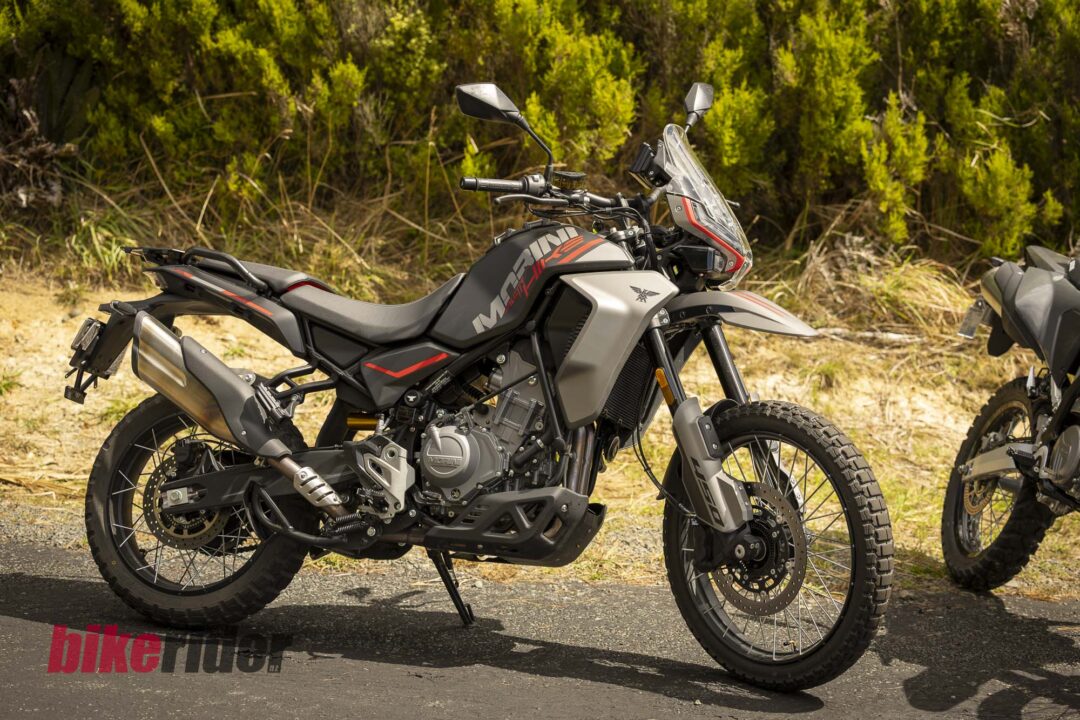 Two adventure motorcycles parked side by side on a paved road with green bushes in the background.
