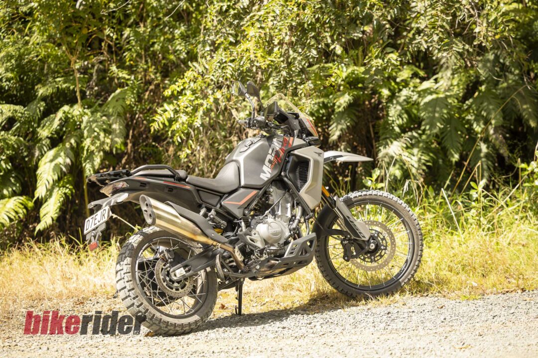 Adventure motorcycle with knobby tires parked on a gravel path beside dense green foliage.