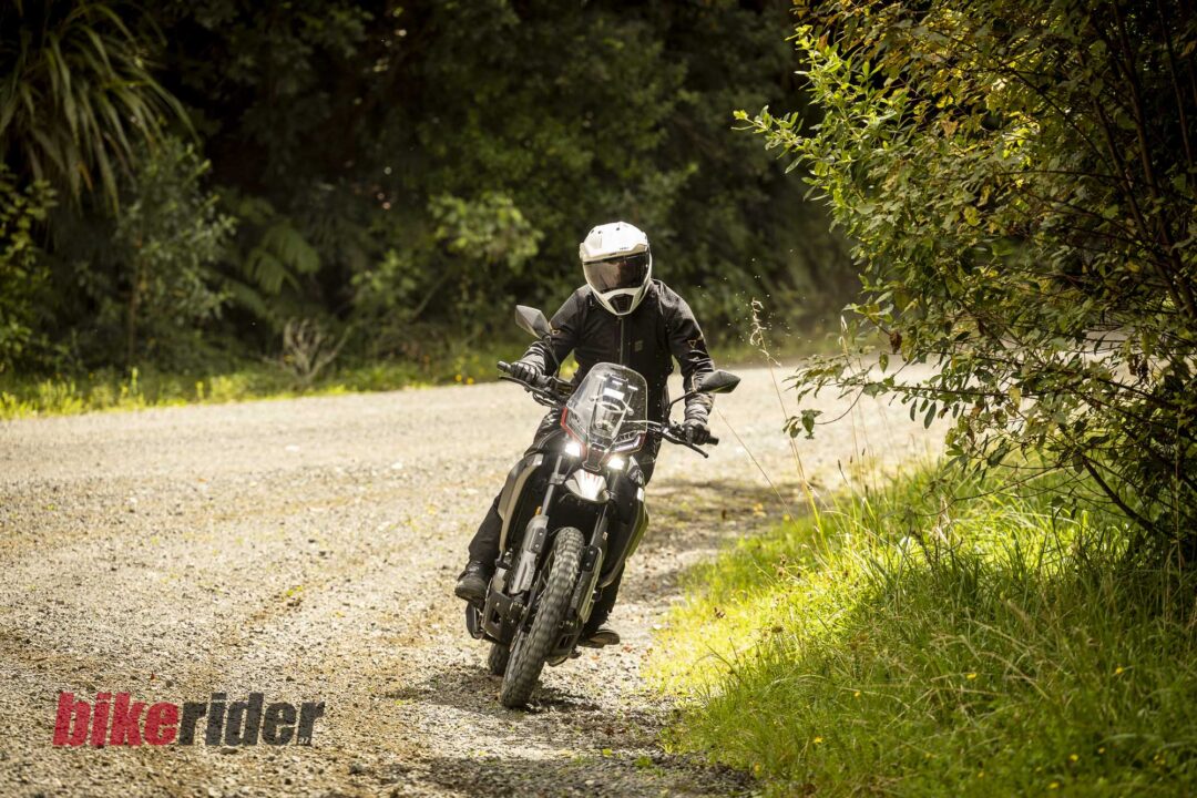 Motorcycle rider in full gear and white helmet leaning into a dirt turn, with greenery on both sides and 'bikerider' logo in the lower-left corner.