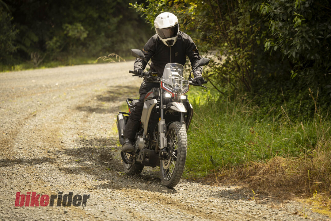 Motorcyclist in protective gear riding a dual-sport motorcycle along a gravel forest road, helmet visor down.