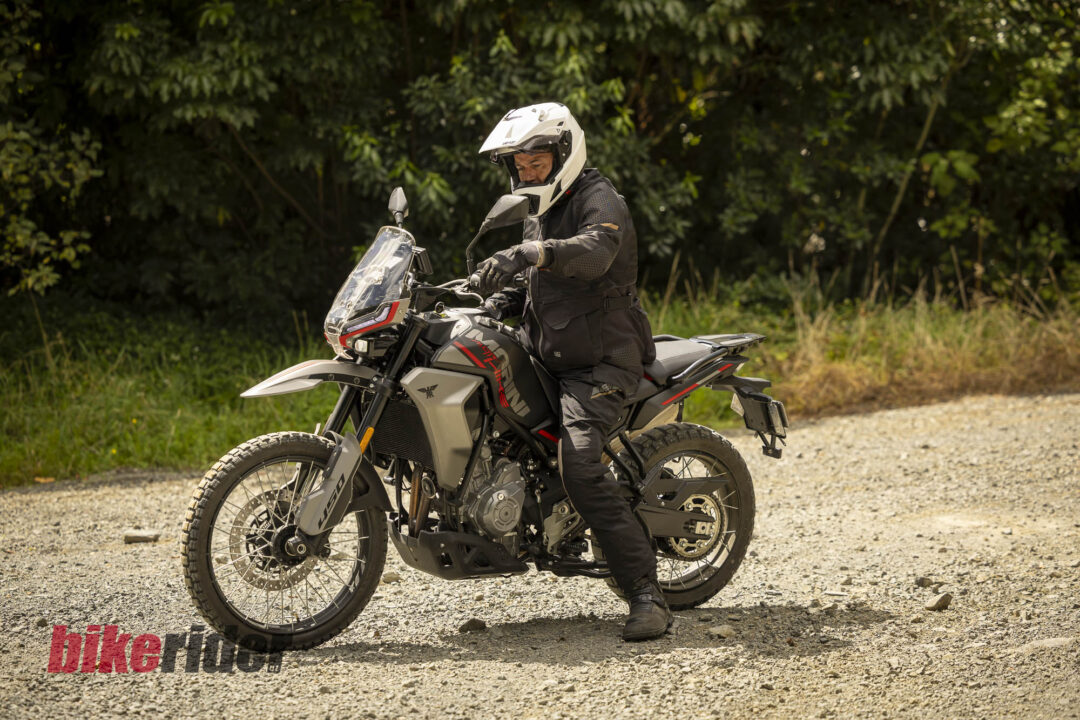 Motorcyclist in a white helmet and protective gear sits on a gray adventure bike on a gravel road with trees behind.