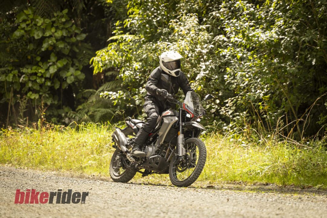 Motocycle rider in full protective gear on a dirt bike in a leafy, sunlit forest setting, riding on a gravel path.