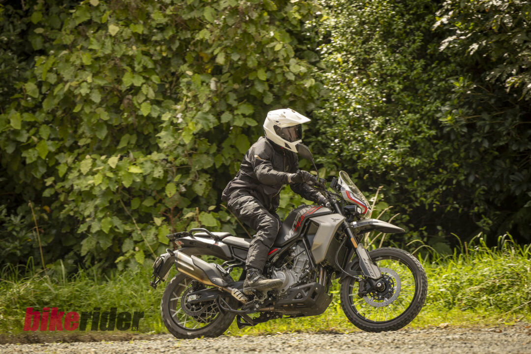 Motorcyclist in full protective gear riding a black and red adventure bike along a gravel path with dense green foliage in the background.