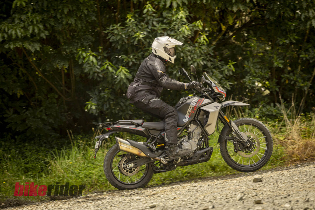 Motorcyclist in a white helmet and black protective gear riding a dual-sport motorcycle along a gravel path with dense green brush in the background.