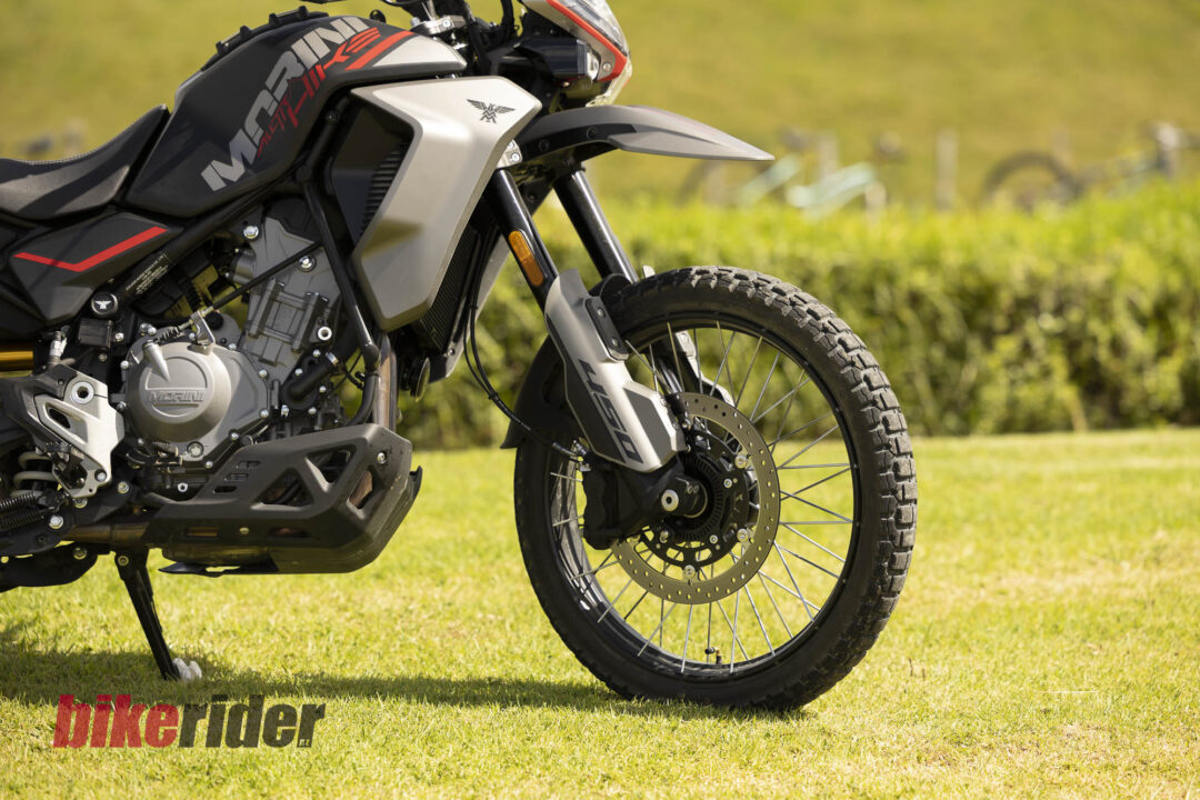 Close-up of a modern dual-sport motorcycle on a grassy field, showing the front wheel, brake disc, and engine details; 'bike rider' text visible on the grass at the bottom left.