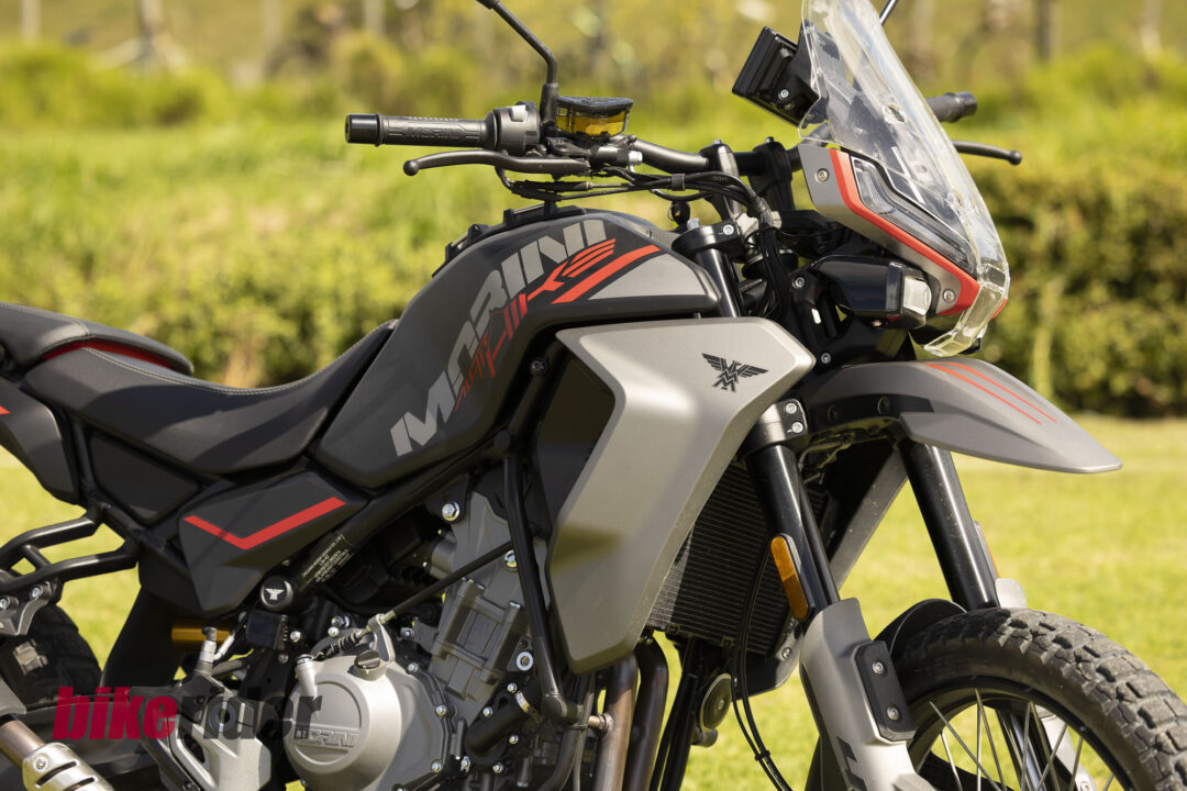 Close-up of a gray and red adventure motorcycle focusing on the handlebar, windshield, and fuel tank with bold graphics, set against a grassy outdoor background.