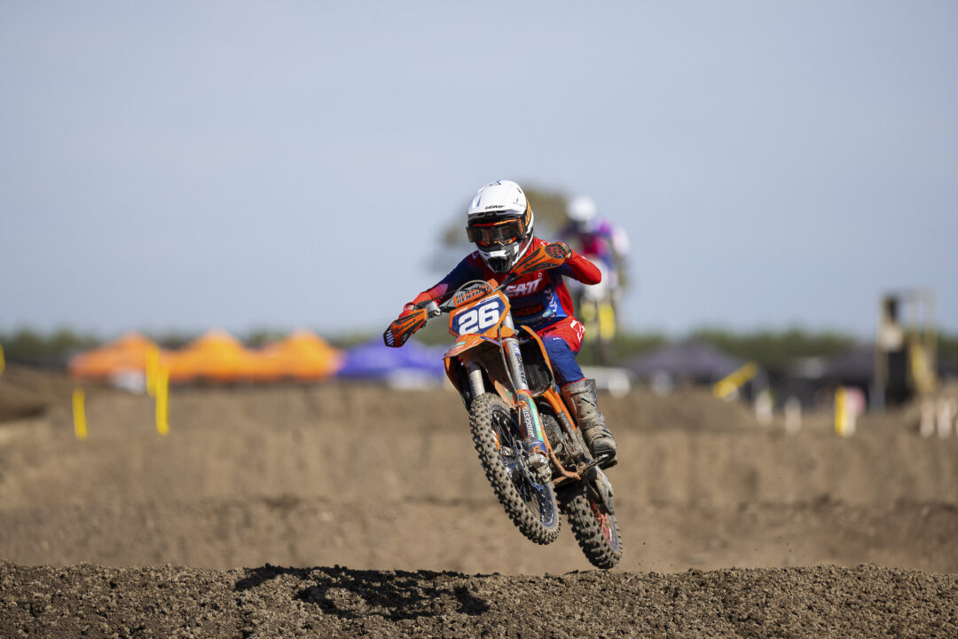 Motocross rider in red and blue gear leaps over a dirt mound on an orange bike, wearing a white helmet and goggles, race scene in background.