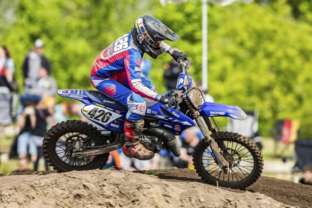 Motocross rider in colorful gear and helmet jumping a dirt bike over a track, spectators in the background.