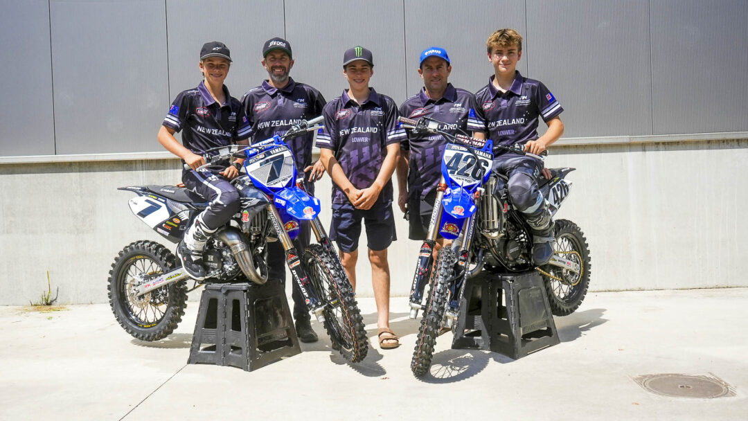 Five motocross riders in matching black‑purple New Zealand jerseys pose with two blue Yamaha dirt bikes on stands outside a gray wall.