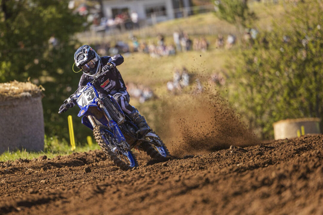 Motorcross rider in black gear leaning into a turn on a dusty dirt track with spectators watching in the background, bike kicking up soil.
