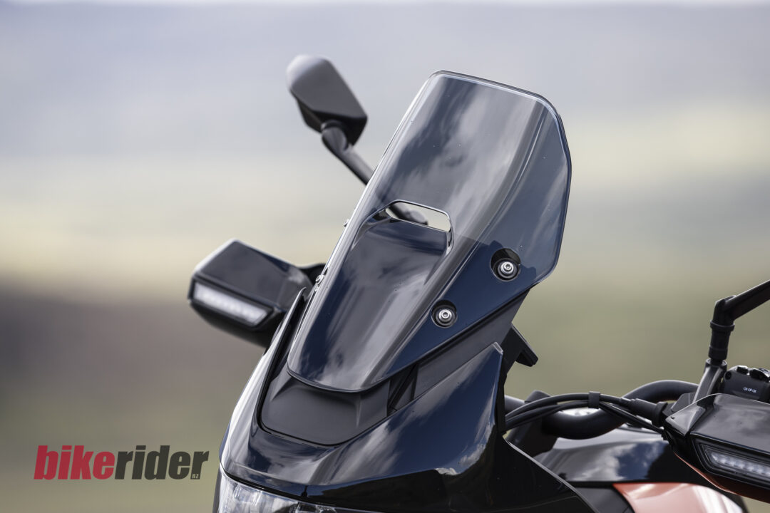 Close-up of a glossy motorcycle windshield and handlebars with a blurred landscape in the background; 'bikerider.nz' logo at bottom left.