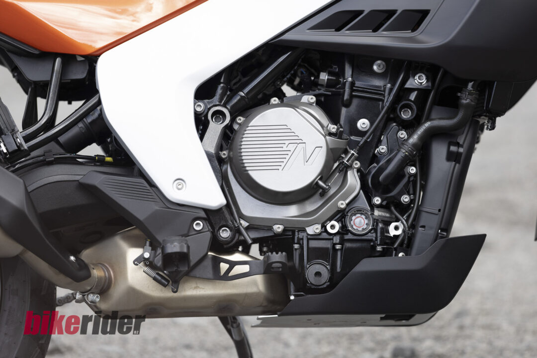 Close-up of Norton Atlas 585cc motorcycle engine beneath white fairing and orange bodywork, showing bolts, hoses, and the metallic engine cover. Watermark 'bikerider.nl' visible at bottom.