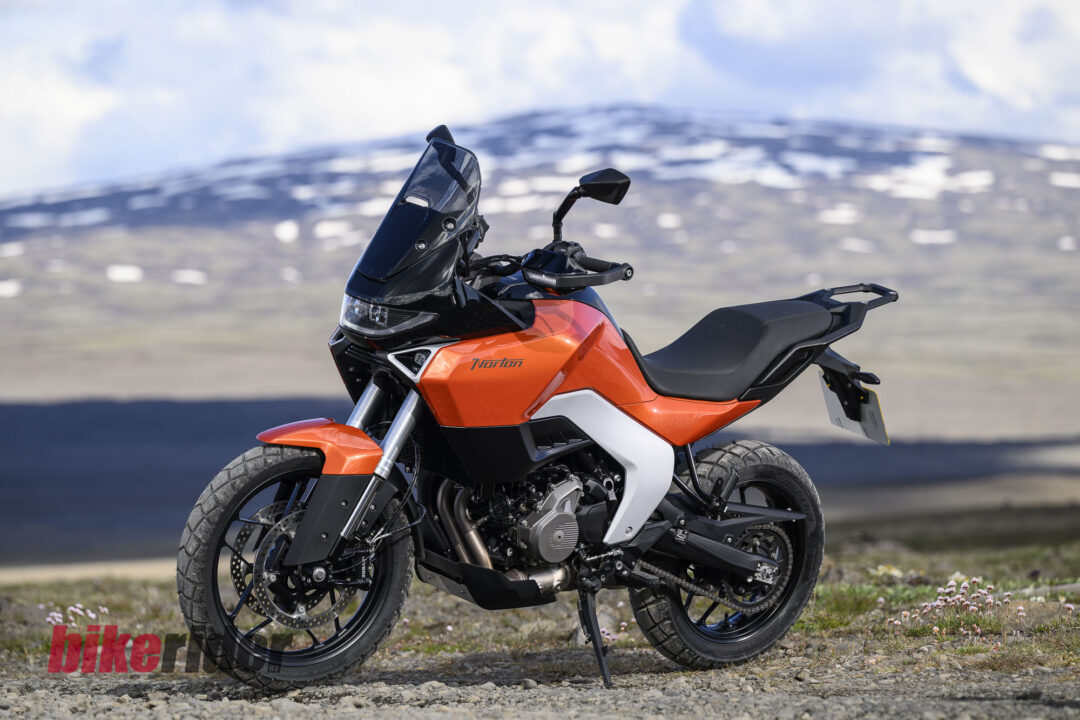 20260703-Norton_Atlas_2026_Offroad_Statics11-1080x720 Orange Norton adventure motorcycle on a rocky dirt path with mountains and blue sky in the background, on its kickstand.
