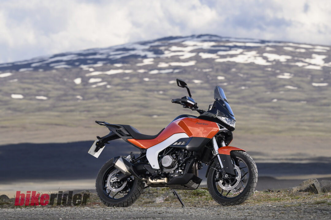 20260703-Norton_Atlas_2026_Offroad_Statics13-1080x720 Orange adventure motorcycle parked on a rocky roadside with snow-dotted hills in the background
