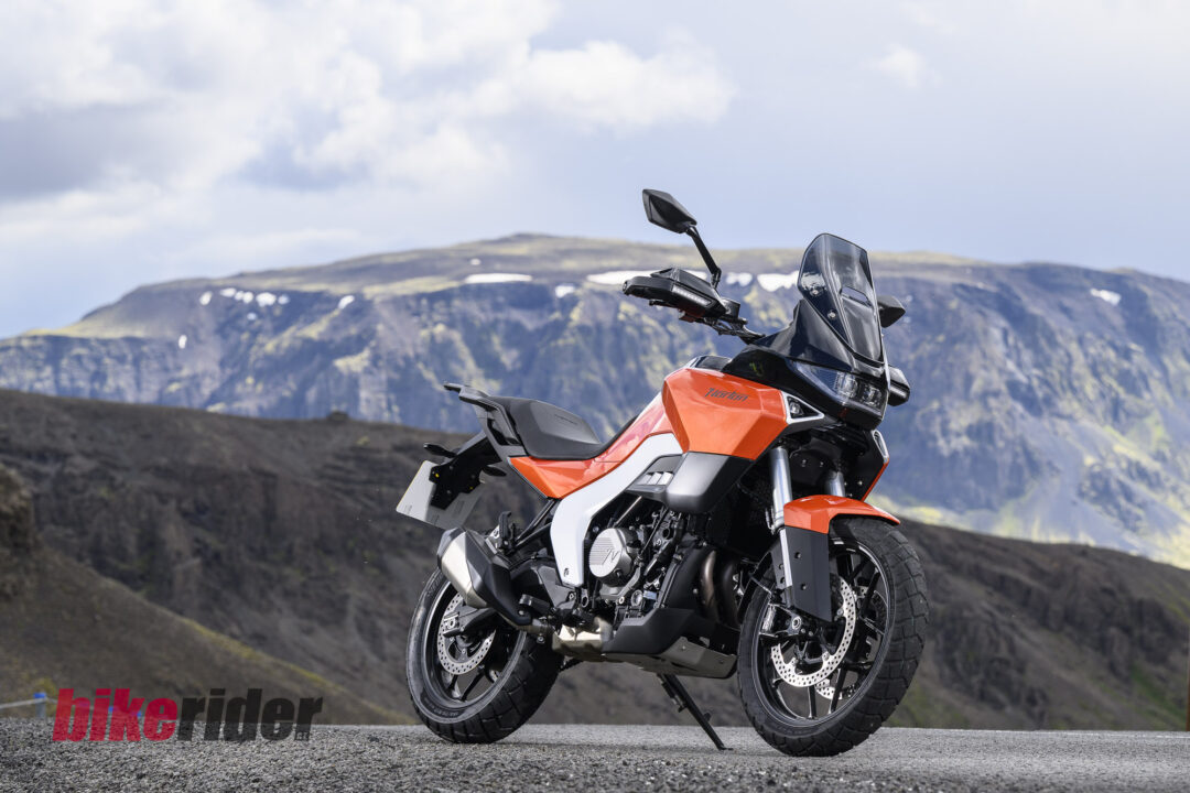 Norton Atlas adventure motorcycle on a mountain road with rocky slopes and a cloudy sky in the background.