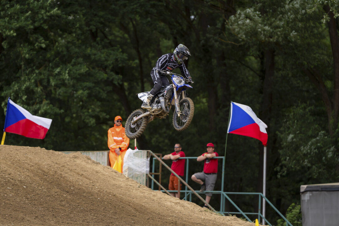 Motocross rider leaps off a dirt ramp during a race, mud-splattered bike in mid-air with a wooded backdrop.