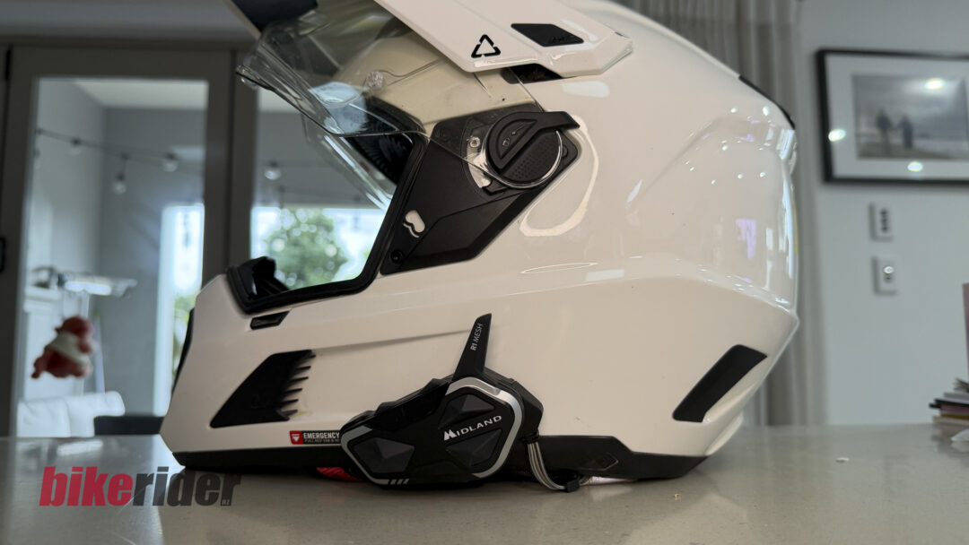 White full-face motorcycle helmet on a showroom counter, visor open, with a black strap and subtle 'bikerider.nz' watermark.