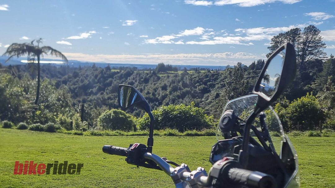 Close-up of a motorcycle handlebar and windshield with a scenic green hillside and distant ocean under a blue sky.