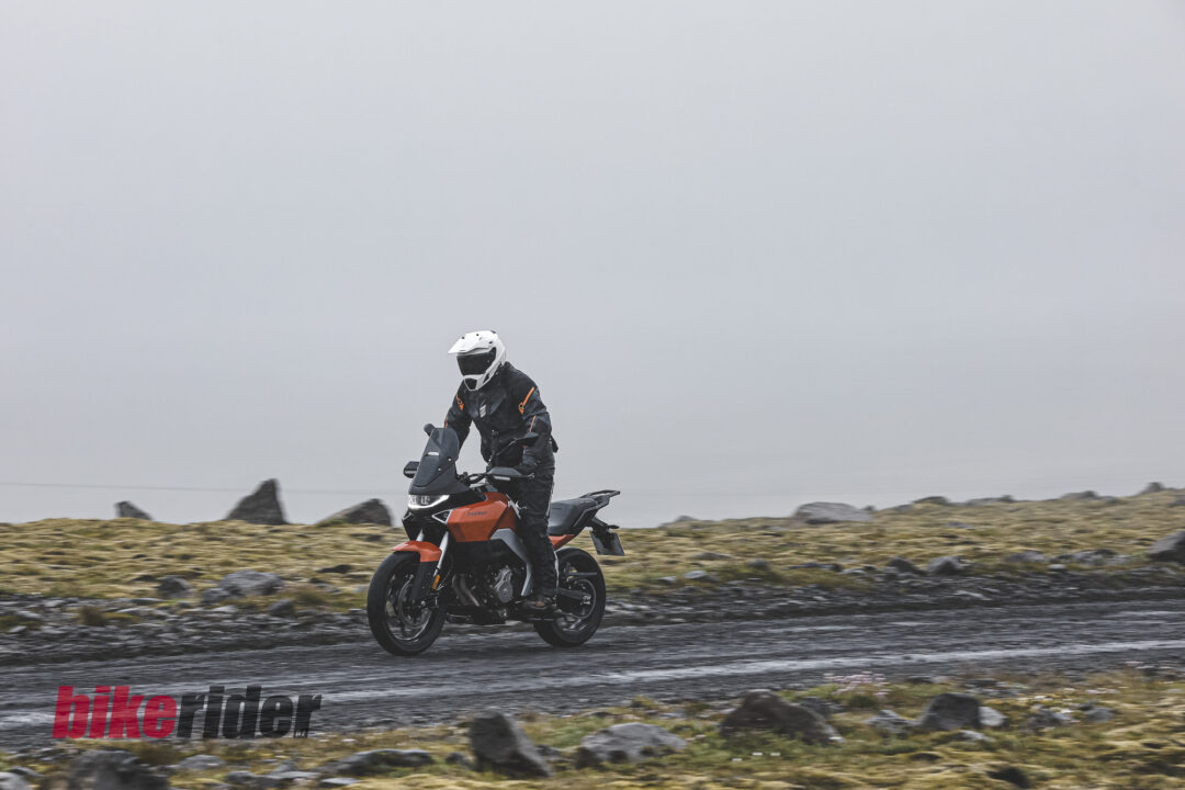 20260712-Norton-Atlas-Global_B_19001-25-1080x720 Motorcyclist wearing a white helmet and protective gear rides a red adventure bike along a rocky coastal road under a cloudy sky.