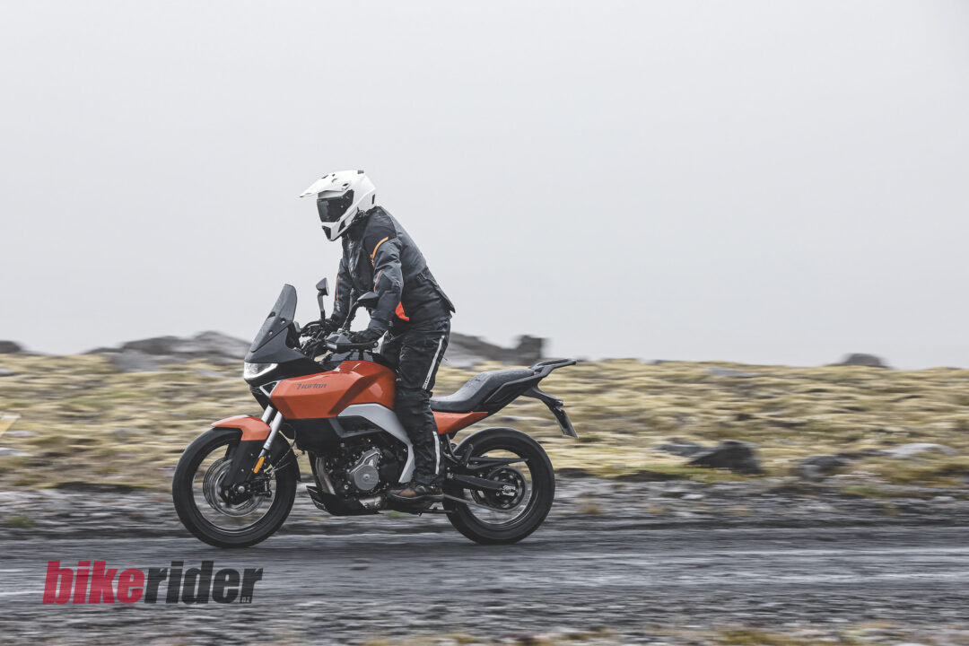 Motorcyclist in a white helmet and dark gear rides an orange adventure bike along a wet, rocky road.